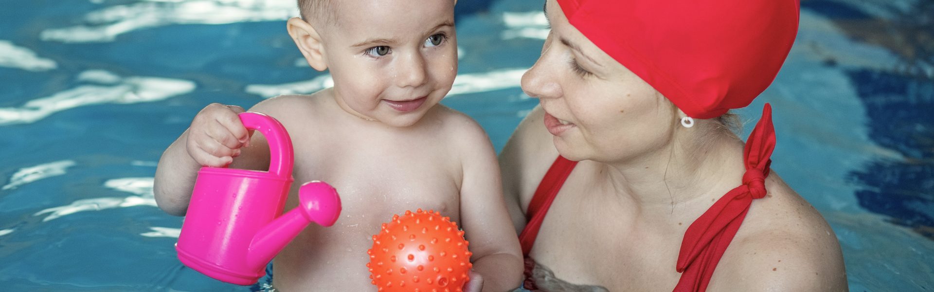 Image shows a parent and toddler playing with a plastic watering can and ball in a pool.